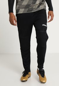Zwarte sportbroek met gestructureerde padding op de knieën, gecombineerd met gouden voetbalschoenen. Heeft een getailleerd ontwerp en een logo op de dij.