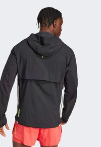 Chaqueta atlética negra con capucha, acentos en un verde claro a contraste y rayas blancas en los lados. Presenta un diseño de espalda en capas y un tejido texturizado.