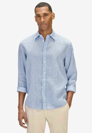 Mand iført en lyseblå skjorte med lange ærmer og mørke lodrette striber samt beige bukser, stående med den ene hånd i lommen.