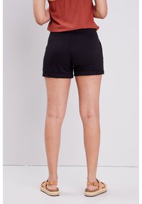 Shorts en coton noir avec des ourlets roulés, dotés de poches arrière. Portés avec un haut couleur rouille et des sandales beiges.