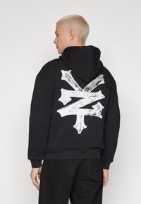 Svart hettegenser med et stort hvitt penselstrøk-motiv på baksiden. Stoffet virker mykt med ribbestrikkede mansjetter og nederkant.