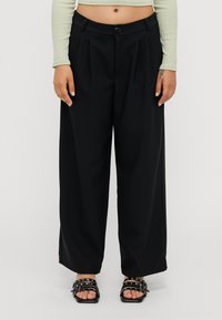 Black wide-leg trousers with pleats, featuring a button closure and a smooth fabric. Worn with black strappy sandals.