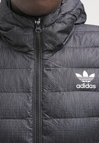 Svart vadderad jacka med en texturerad tyg, hög krage och dragkedja. Har en vit Adidas-logotyp på bröstet.