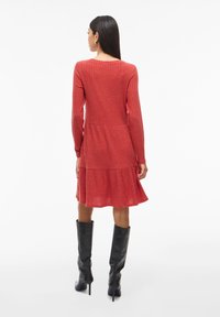 Červené žebrované šaty s dlouhými rukávy, vrstvenou sukní a zakulaceným výstřihem. V kombinaci s vysokými černými botami. Hladká textura.