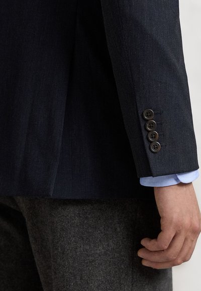 Manche de blazer bleu foncé avec une texture lisse, quatre boutons marron, et une manche de chemise bleu clair visible en dessous. Pantalon gris partiellement visible.
