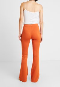 Pantalons évasés orange en tissu extensible, avec une taille haute et une texture lisse, vus de dos avec un débardeur blanc.