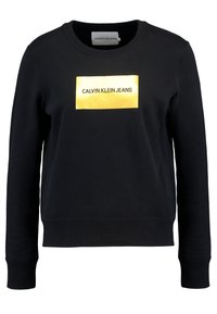 Černá mikina s kulatým výstřihem a dlouhými rukávy, na které se nachází zlatý obdélníkový logo se textem "CALVIN KLEIN JEANS" uprostřed.