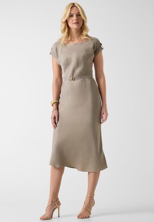 Kobieta ubrana w taupe midi sukienkę z paskiem i krótkimi rękawami, złote bransoletki oraz złote sandały na wysokim obcasie z paskami, stojąca na białym tle.