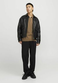 Veste en cuir noir avec un devant zippé, polo marron en dessous, pantalon noir ample et chaussures noires. Design minimaliste.