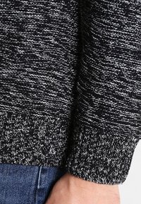 Czarnobiały melanżowy mankiet swetra z dzianiny na nadgarstku i dłoni osoby, na tle niebieskich dżinsów.