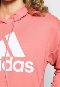 Sweat à capuche rose clair en tissu doux, avec un grand logo Adidas blanc sur la poitrine et une capuche avec cordon.