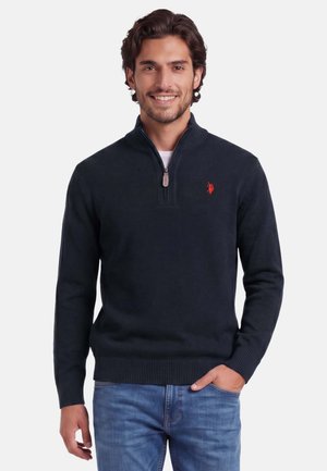 Lächelnder Mann mit dunklen Haaren, der einen marineblauen Viertel-Reißverschluss-Pullover mit rotem Logo und blauen Jeans trägt, Hand in der Tasche, vor weißem Hintergrund.