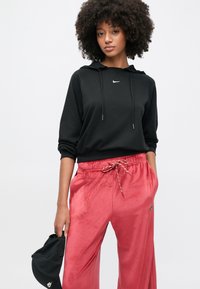 Žena s kudrnatými vlasy, oblečená v černé mikině Nike a červených sametových kalhotách, držící v levé ruce černou čepici Nike na jednobarevném pozadí.