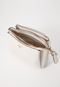 Helle, strukturierte Tasche in Cremefarbe mit goldenen Reißverschlüssen, ausgestattet mit drei Fächern und schwarzen Lederelementen. Logodetail auf der Vorderseite.