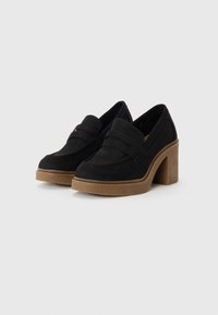 Mocassins en daim noir à bout carré, dotés d'une sangle décorative et d'un talon épais et texturé marron. La semelle est légèrement surélevée.