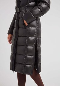 Abrigo de plumas negro con cintura ceñida, que presenta un patrón acolchado, bolsillos laterales y una longitud hasta la mitad de la pantorrilla, con un material brillante y suave.