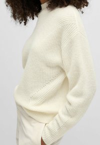 Femme portant un pull en tricot côtelé blanc cassé à manches longues, associé à un pantalon clair, main dans la poche, cheveux bouclés visibles.