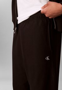 Osoba poprawiająca białą koszulę wpuszczoną do czarnych spodni dresowych Calvin Klein z zamkiem, w czarnej kurtce z widocznym suwakiem.