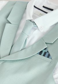 Mint zelen jopič s pripadajočim kravatnim trakom, pod katerim je bela srajca z ovratnikom. Opaženi vzorci v žepu jopiča. Gladek tekstura, krojena zasnova.