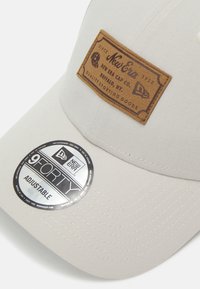 Adjustable beige cap with a textured brown leather patch labeled "New Era." Features a silver circular tag stating "9FORTY."