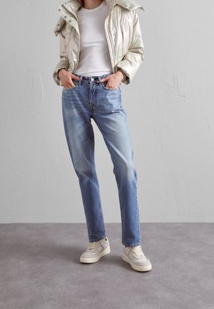 Persona vestida con una chaqueta de plumas color crema, camiseta blanca de canalé, jeans de un azul claro y zapatillas blancas, de pie contra una pared y un suelo gris liso.