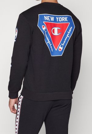 Persoon draagt een zwarte trui en broek met Champion-logo en de tekst "New York" op de achterkant, en logostrepen langs de zijkanten van de broek.