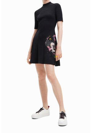 Personne portant une robe noire à manches courtes avec un motif coloré de Mickey Mouse et des baskets blanches, debout devant un fond blanc.