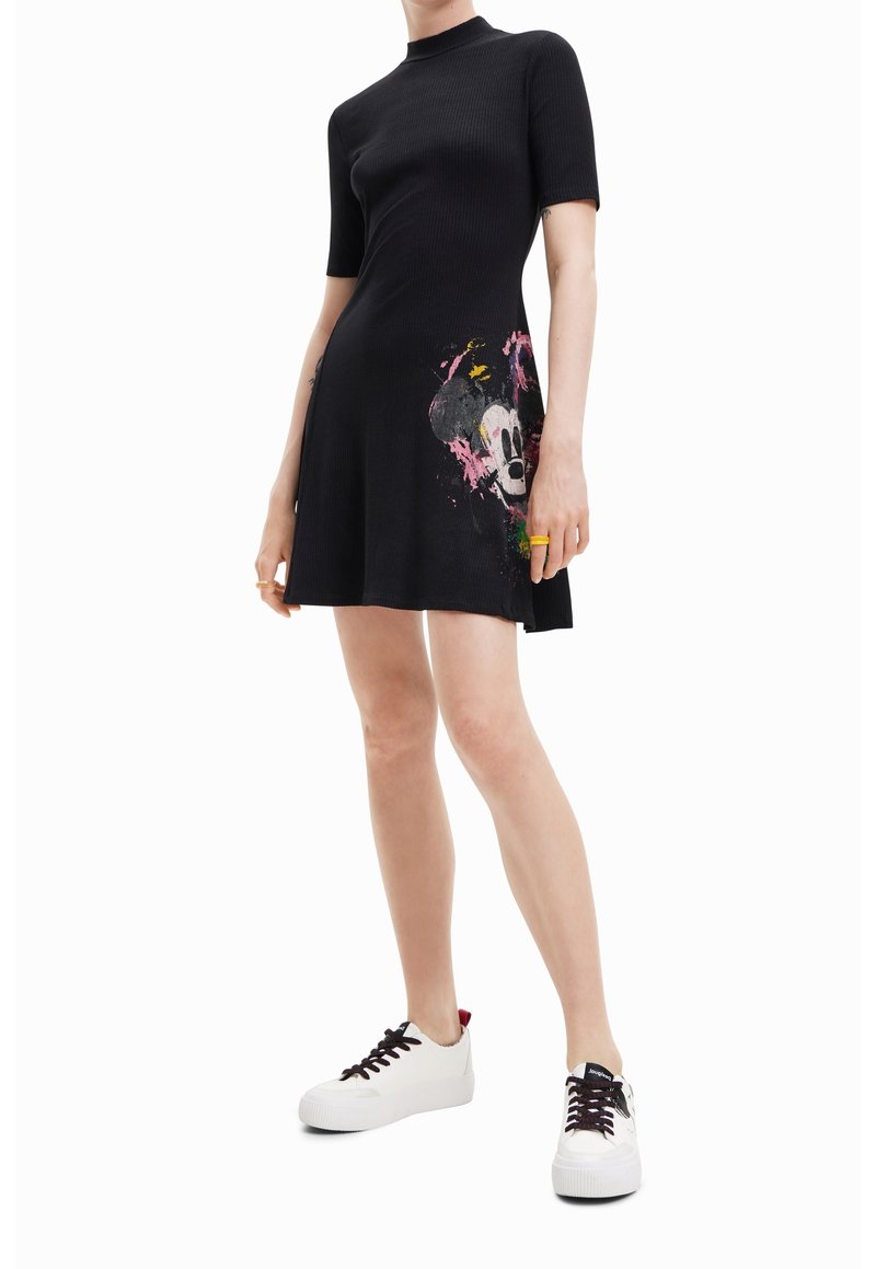 Personne portant une robe noire à manches courtes avec un motif coloré de Mickey Mouse et des baskets blanches, debout devant un fond blanc.