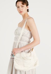 Witte stoffen schoudertas met een geplooide textuur en twee handvatten bovenaan. De tas heeft een enkele verstelbare schouderriem voor crossbody gebruik.