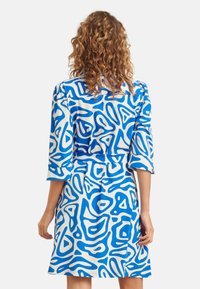 Kurzes ärmelloses Kleid mit tailliertem Gürtel, das ein abstraktes blau-weißes Muster auf einem glatten Stoff zeigt, in mittlerer Oberschenkellänge.