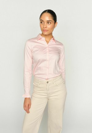 Frau, die leicht nach links blickt, trägt eine hellrosa Bluse mit langen Ärmeln und Rüschenbündchen sowie eine cremefarbene, hoch taillierte Hose mit Nietenverzierung.