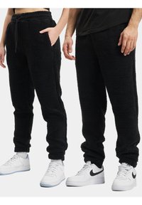 Sorte fleece joggere med en myk tekstur, elastisk midje og mansjetter. Side-lommer synlige på venstre bukse. Brukt med hvite sneakers.