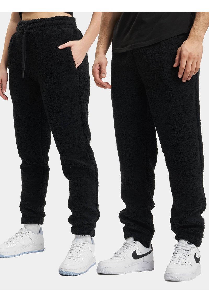 Sorte fleece joggere med en myk tekstur, elastisk midje og mansjetter. Side-lommer synlige på venstre bukse. Brukt med hvite sneakers.