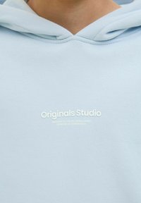 Világoskék kapucnis pulóver, puha anyagból készült, a közepén a "Originals Studio" felirat fehér színben látható, elöl zsebbel és húzózsinórral ellátva.
