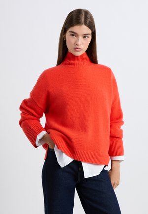 Maglione arancione brillante a collo alto oversize realizzato in tessuto morbido, con polsini e orlo a costine, indossato sopra una camicia bianca e jeans scuri.