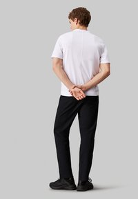 Camicia bianca a maniche corte, pantaloni neri e scarpe nere; vista da dietro, con le mani intrecciate dietro la schiena. Design semplice e forma aderente.