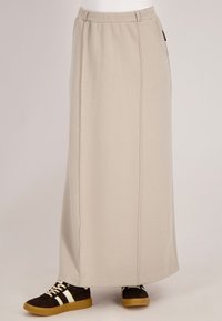 Beige maxi rok van zachte stof, met een tailleband en zijzakken. Gecombineerd met bruine sneakers met witte strepen.