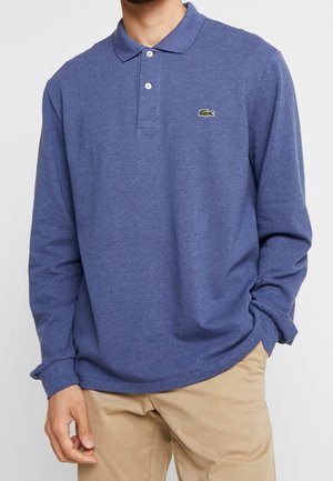 Homme portant un polo bleu à manches longues avec deux boutons et un pantalon beige, debout avec une main dans la poche.