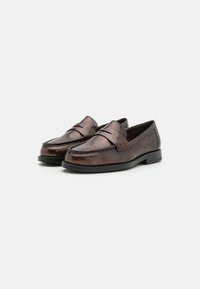 Mocassins de couro castanho com um acabamento texturizado, apresentando um detalhe de tira elevada e uma ponta arredondada, combinados com uma sola preta em contraste.