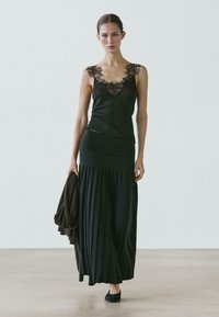 Donna che indossa una canottiera nera con bordo in pizzo e una lunga gonna plissettata, che tiene in mano un cappotto scuro, camminando su un pavimento neutro davanti a una parete semplice.