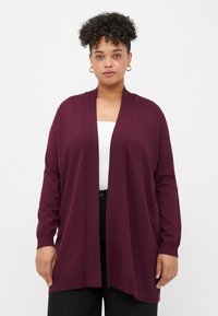 Woman with curly hair wearing gold hoop earrings, a white top, black pants, and a loose, open-front burgundy cardigan.