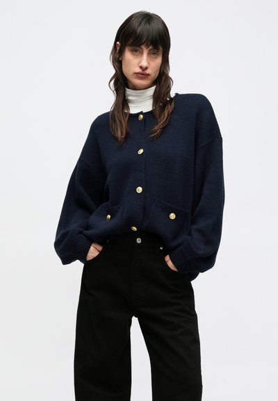 Tmavomodrý pletený sveter s nadrozmerným strihom, ozdobený zlatými gombíkmi, dvoma prednými vreckami a rebrovanými manžetami, vrstvený cez biely rolák.