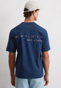 Granatowa koszulka z krótkim rękawem, z nadrukowanymi współrzędnymi i napisem "marc o'polo" w białym kolorze na plecach. Wykonana z miękkiej bawełny.
