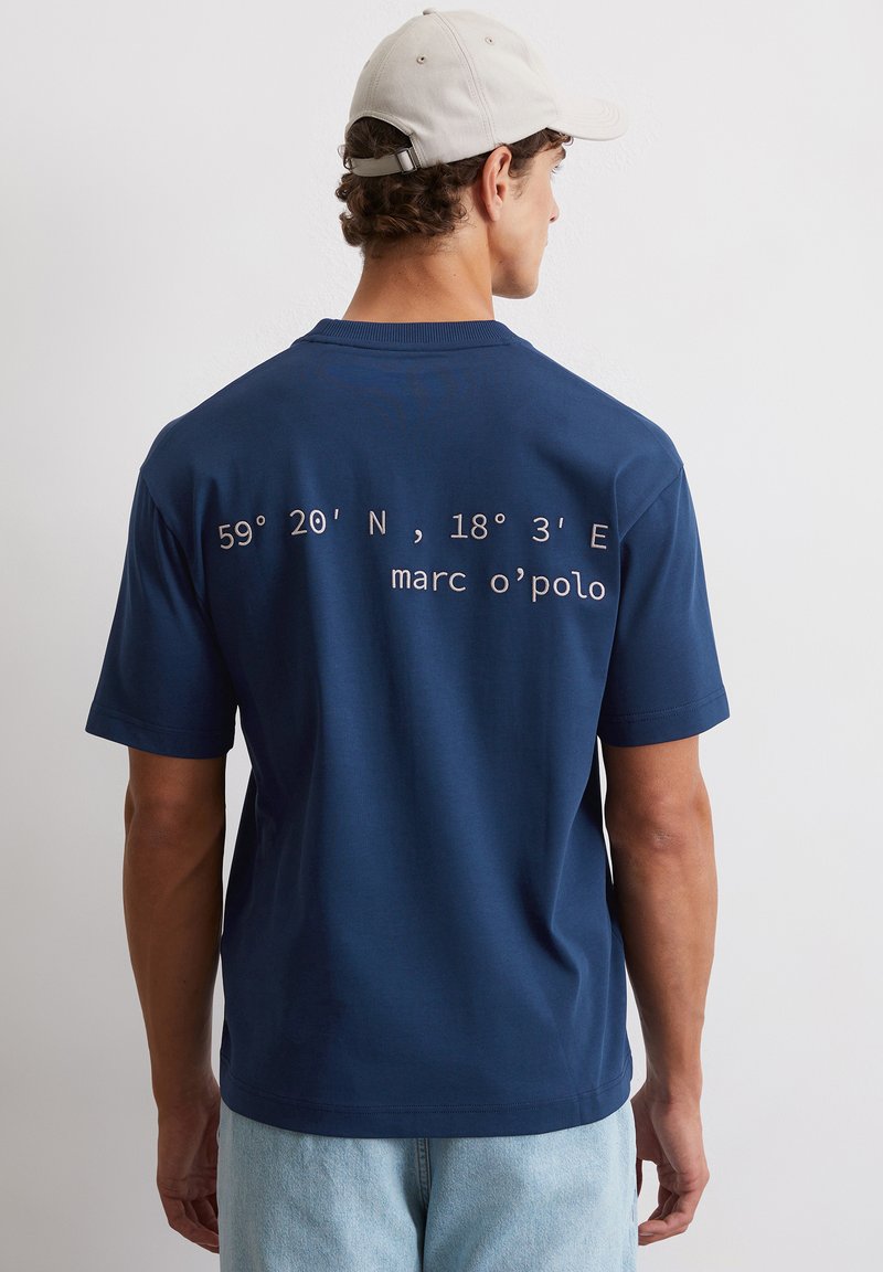 Granatowa koszulka z krótkim rękawem, z nadrukowanymi współrzędnymi i napisem "marc o'polo" w białym kolorze na plecach. Wykonana z miękkiej bawełny.