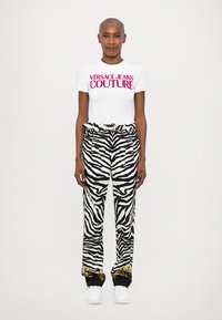 Balta kreklisa ar rozā "VERSACE JEANS COUTURE" tekstu, kombinēta ar augstām melnbaltām zebras svītrām, kurām ir zelta akcentēti aproces.
