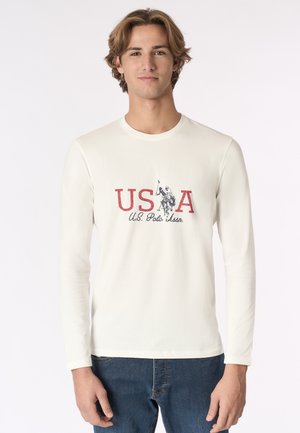 Camicia a maniche lunghe color crema con una grafica rossa "USA" e il logo blu della U.S. Polo Association. Il tessuto appare morbido e leggero.