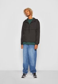 Sort letvægts hættetrøje med lynlås fortil, parret med en grøn skjorte og løstsiddende blå jeans, båret med sorte og hvide sneakers.
