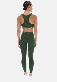 Tamsiai žalia sportinė liemenėlė ir tamprės komplektas pasižymi švelnia tekstūra, patogiu prigludimu ir prekiniu elastiniu liemeniu. Derinamas su sportiniais bateliais.