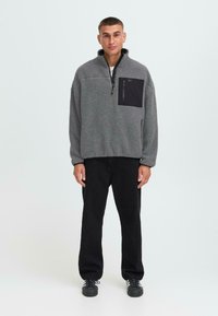 Fleece-Pullover in Hellgrau mit Reißverschlusskragen und schwarzer Brusttasche; strukturiertes Material; kombiniert mit schwarzen Hosen und dunklen Schuhen.
