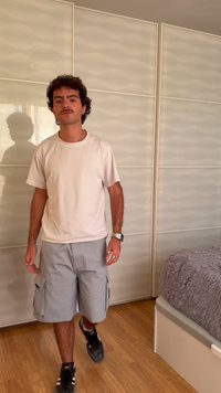 Νέος άνδρας με σγουρά μαλλιά και μουστάκι περπατάει σε εσωτερικό χώρο φορώντας λευκή μπλούζα, γκρι cargo σορτς, μαύρα αθλητικά παπούτσια και ρολόι χειρός.